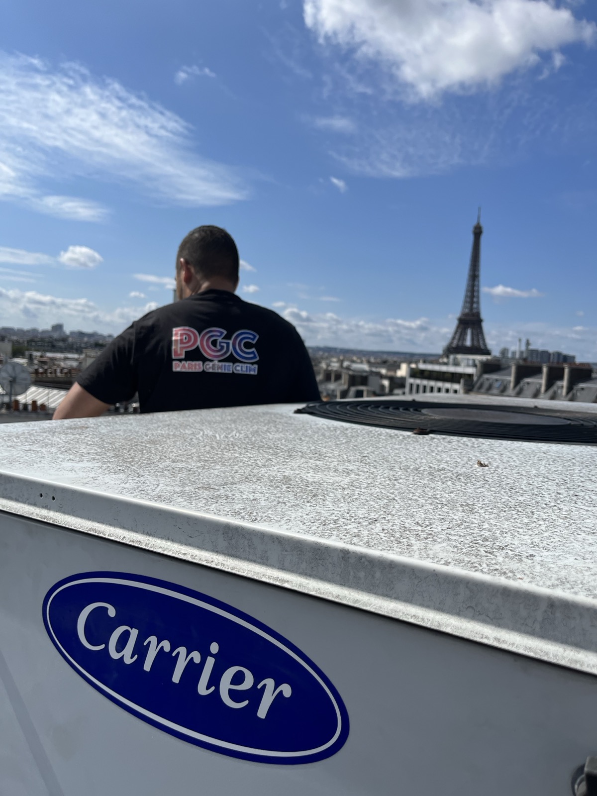 Un technicien Paris Génie Clim, vu de dos, en intervention sur un toit parisien ; au premier plan le capot d'un groupe de climatisation Carrier, la tour Eiffel en arrière-plan.