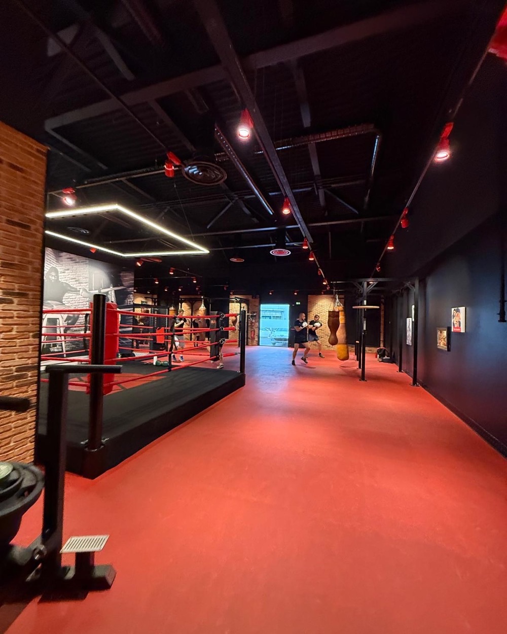 Espace boxe d'On Air Fitness : ring et sacs de frappe sur sol rouge, diffuseurs d'air et gaine apparents sous la charpente noire.