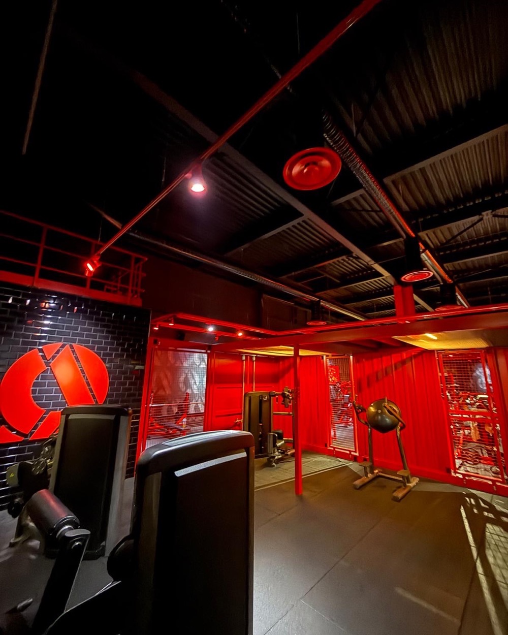 Zone d'entraînement d'On Air Fitness : diffuseurs d'air circulaires rouges et gaines suspendus au plafond noir, au-dessus d'un espace aménagé en conteneur rouge.