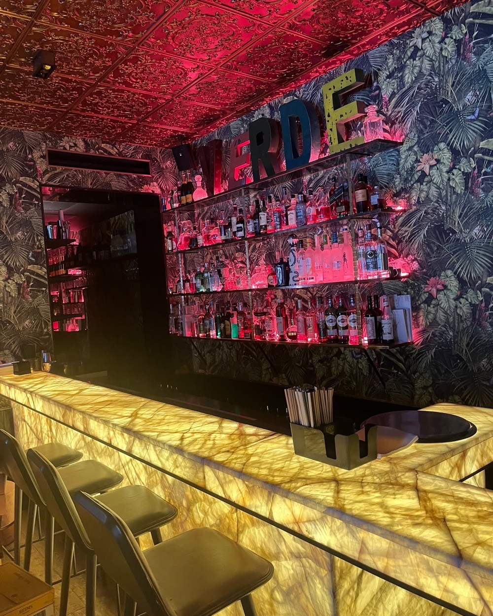 Bar du restaurant Verde : comptoir lumineux, étagères de bouteilles et grille de soufflage linéaire au plafond.